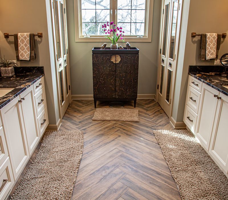 Bronze crosshatched mirror frames, herringbone wood look floor tile, and a vaulted ceiling add distinction to this two vanity bathroom.