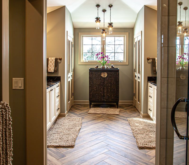 Bronze crosshatched mirror frames, herringbone wood look floor tile, and a vaulted ceiling add distinction to this two vanity bathroom.