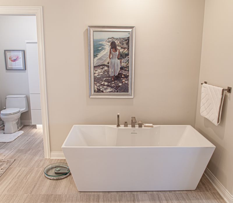 The neutral colors of the chevron wall tiles, spacious tiled walk-in shower, minimalist cabinetry, and a modern freestanding tub all set off the painting that is the centerpiece of this bathroom.