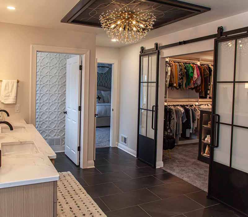 Elegance reigns supreme in this bathroom remodel that features an en suite, walk-in closet, walk-in shower, custom double vanity, private water closet, framed-glass barn doors, and a dazzling starburst chandelier.