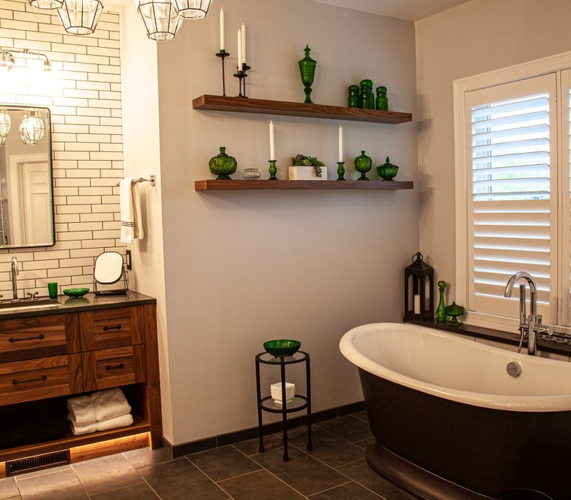 A retro inspired bathroom with a black and white freestanding tub, green glass accents, white subway tiles with dark grout, chrome fixtures, and walnut cabinetry.