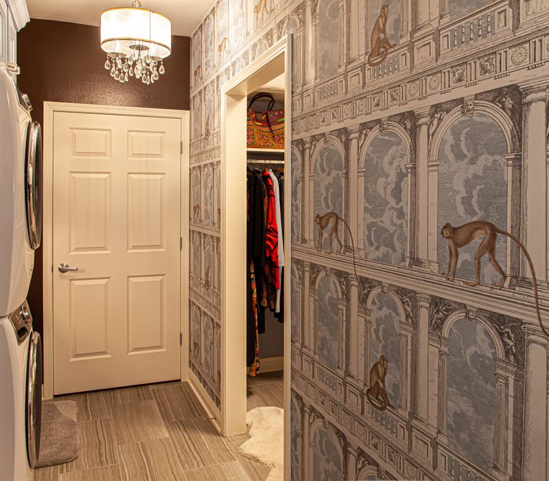 Wallpaper featuring monkeys posing on Venetian architecture, light blue cabinets, and elongated hexagon mosaic tiles add interest to this elegant bathroom with laundry machines and an attached walk-in closet.
