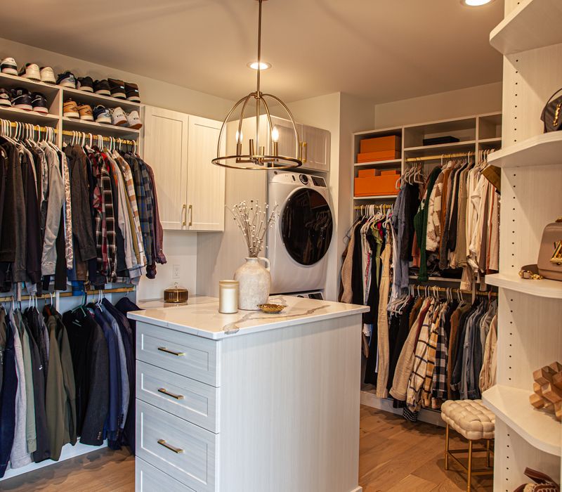 White cabinetry and shelving, a wood floor, and a gold chandelier add beauty to this functional walk-in closet that includes a washer and dryer.