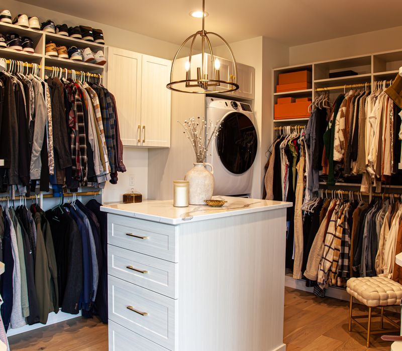 White cabinetry and shelving, a wood floor, and a gold chandelier add beauty to this functional walk-in closet that includes a washer and dryer.