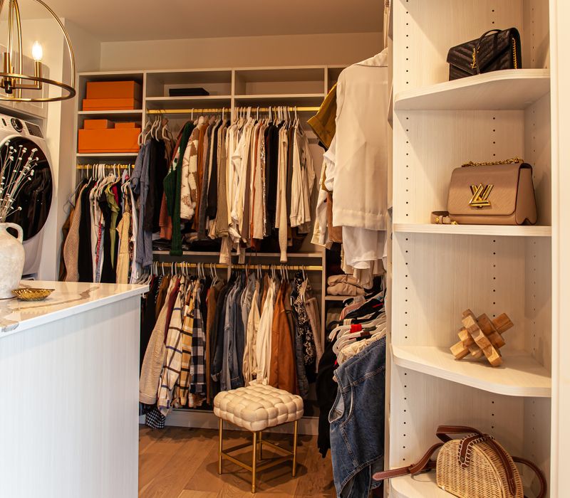 White cabinetry and shelving, a wood floor, and a gold chandelier add beauty to this functional walk-in closet that includes a washer and dryer.
