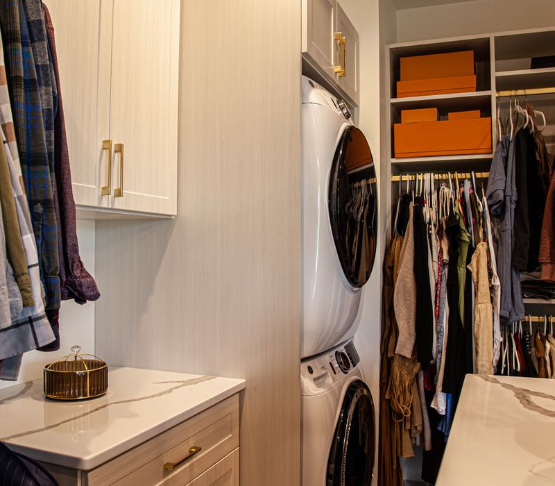 White cabinetry and shelving, a wood floor, and a gold chandelier add beauty to this functional walk-in closet that includes a washer and dryer.