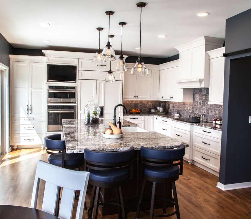 This kitchen combines patterned wall tile, stone countertops, Edison pendant lights, and elegant white cabinetry with a rustic barn door pantry and heavy wooden beams to define the space.