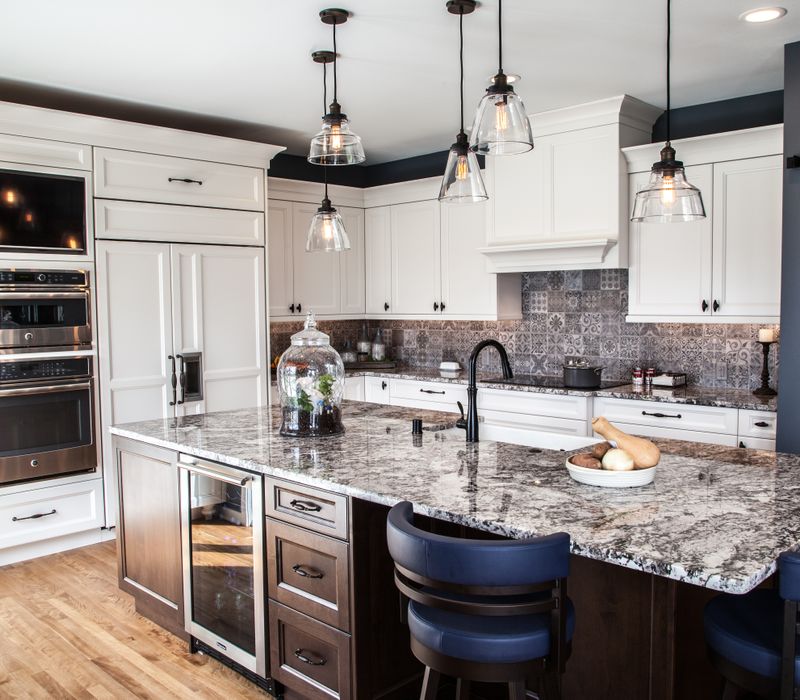 This kitchen combines patterned wall tile, stone countertops, Edison pendant lights, and elegant white cabinetry with a rustic barn door pantry and heavy wooden beams to define the space.