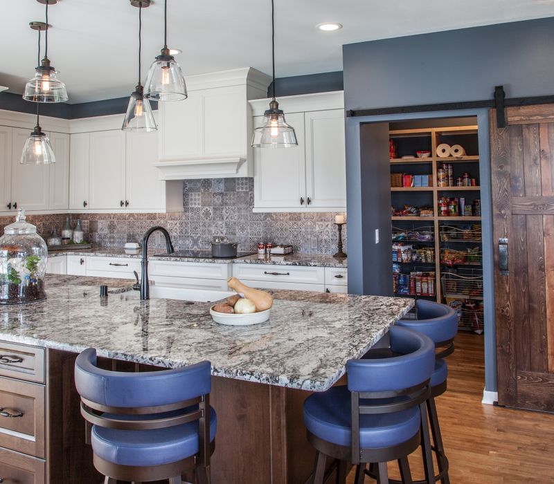 This kitchen combines patterned wall tile, stone countertops, Edison pendant lights, and elegant white cabinetry with a rustic barn door pantry and heavy wooden beams to define the space.
