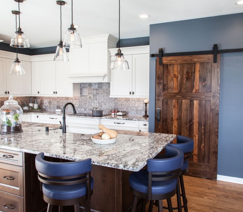 This kitchen combines patterned wall tile, stone countertops, Edison pendant lights, and elegant white cabinetry with a rustic barn door pantry and heavy wooden beams to define the space.