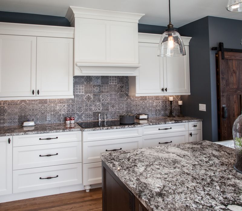 This kitchen combines patterned wall tile, stone countertops, Edison pendant lights, and elegant white cabinetry with a rustic barn door pantry and heavy wooden beams to define the space.