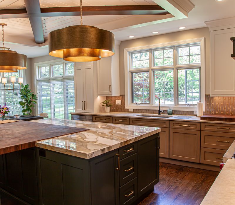 This warm, inviting kitchen features modern brushed gold chandeliers, marble countertops with gold veins, an end-grain cutting board, a metallic glass tile backsplash, and a black range hood with brass accents.