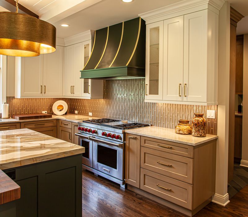 This warm, inviting kitchen features modern brushed gold chandeliers, marble countertops with gold veins, an end-grain cutting board, a metallic glass tile backsplash, and a black range hood with brass accents.