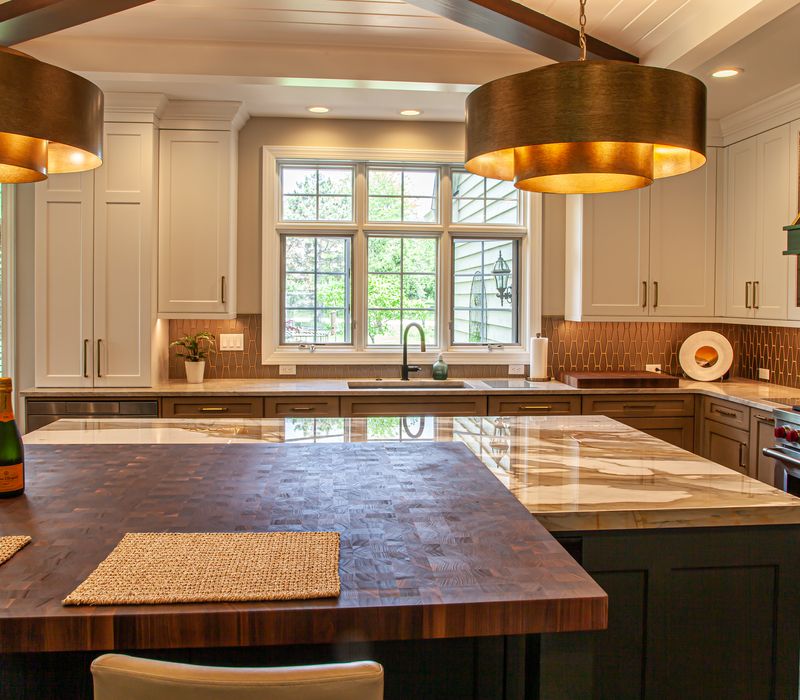 This warm, inviting kitchen features modern brushed gold chandeliers, marble countertops with gold veins, an end-grain cutting board, a metallic glass tile backsplash, and a black range hood with brass accents.