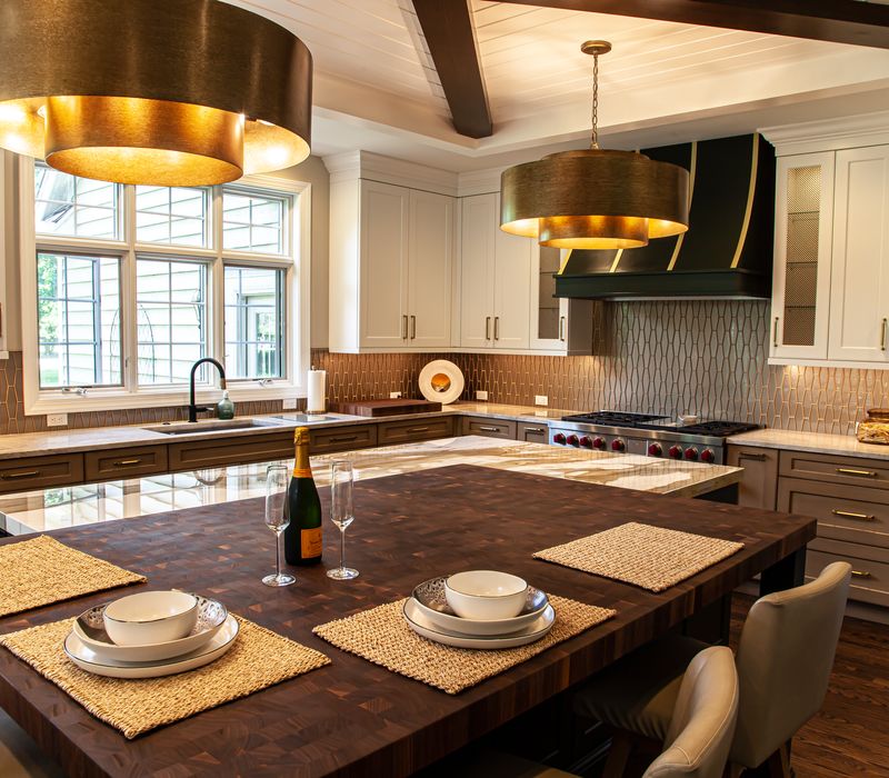 This warm, inviting kitchen features modern brushed gold chandeliers, marble countertops with gold veins, an end-grain cutting board, a metallic glass tile backsplash, and a black range hood with brass accents.