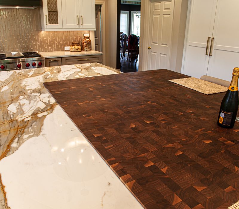 This warm, inviting kitchen features modern brushed gold chandeliers, marble countertops with gold veins, an end-grain cutting board, a metallic glass tile backsplash, and a black range hood with brass accents.
