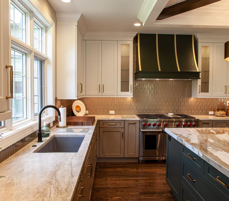 This warm, inviting kitchen features modern brushed gold chandeliers, marble countertops with gold veins, an end-grain cutting board, a metallic glass tile backsplash, and a black range hood with brass accents.