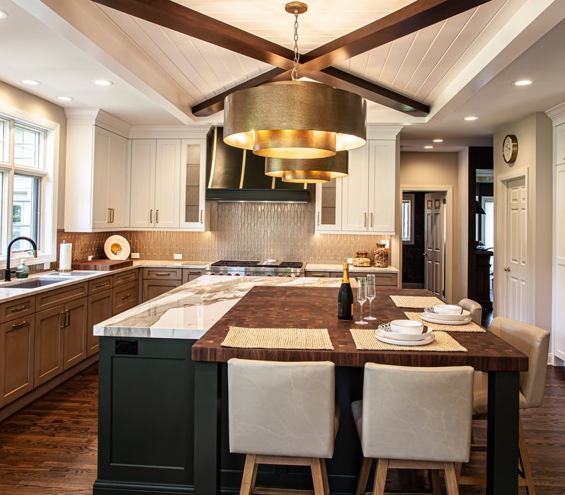 This warm, inviting kitchen features modern brushed gold chandeliers, marble countertops with gold veins, an end-grain cutting board, a metallic glass tile backsplash, and a black range hood with brass accents.