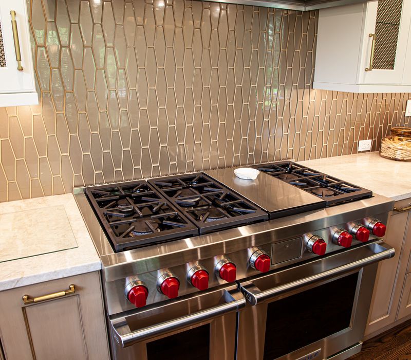 This warm, inviting kitchen features modern brushed gold chandeliers, marble countertops with gold veins, an end-grain cutting board, a metallic glass tile backsplash, and a black range hood with brass accents.