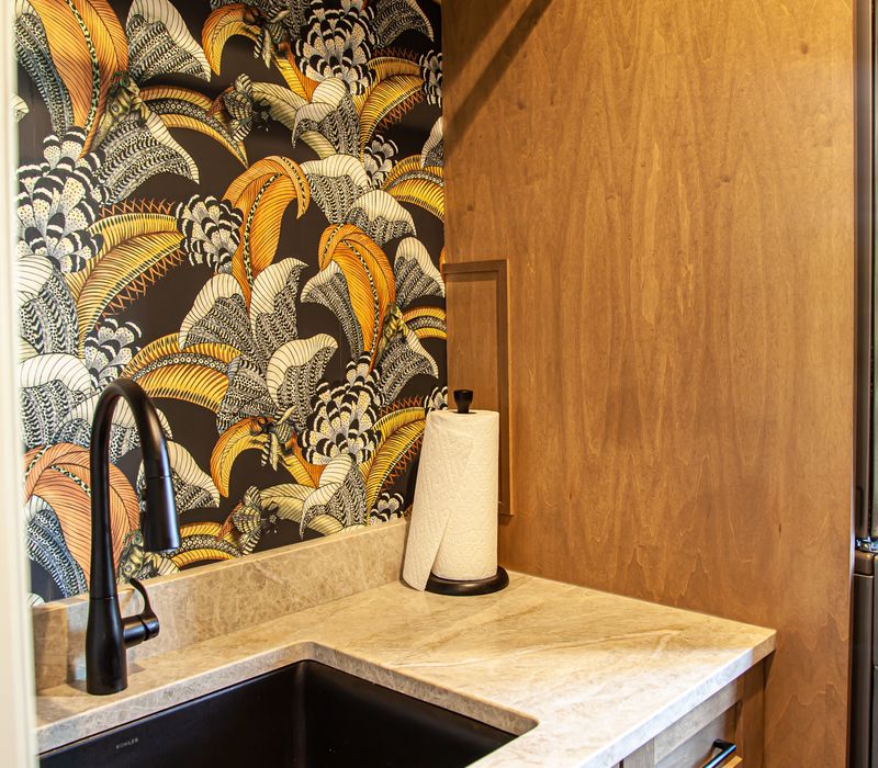 This warm, inviting kitchen features modern brushed gold chandeliers, marble countertops with gold veins, an end-grain cutting board, a metallic glass tile backsplash, and a black range hood with brass accents.