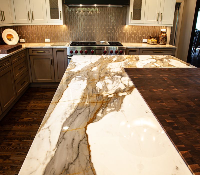 This warm, inviting kitchen features modern brushed gold chandeliers, marble countertops with gold veins, an end-grain cutting board, a metallic glass tile backsplash, and a black range hood with brass accents.