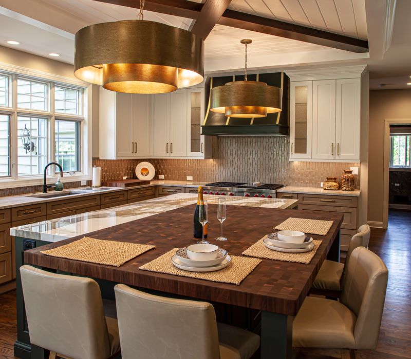 This warm, inviting kitchen features modern brushed gold chandeliers, marble countertops with gold veins, an end-grain cutting board, a metallic glass tile backsplash, and a black range hood with brass accents.