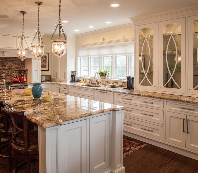 Hardwood floors, polished stone countertops, a mosaic stone tile backsplash, white glass-front cabinetry, and glass pendant lanterns combine in this spacious kitchen.