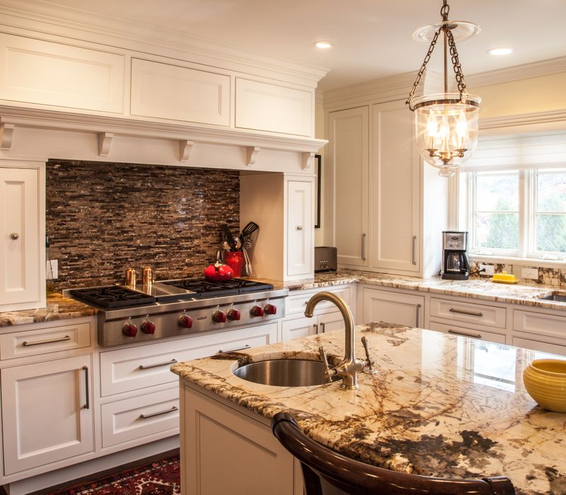 Hardwood floors, polished stone countertops, a mosaic stone tile backsplash, white glass-front cabinetry, and glass pendant lanterns combine in this spacious kitchen.