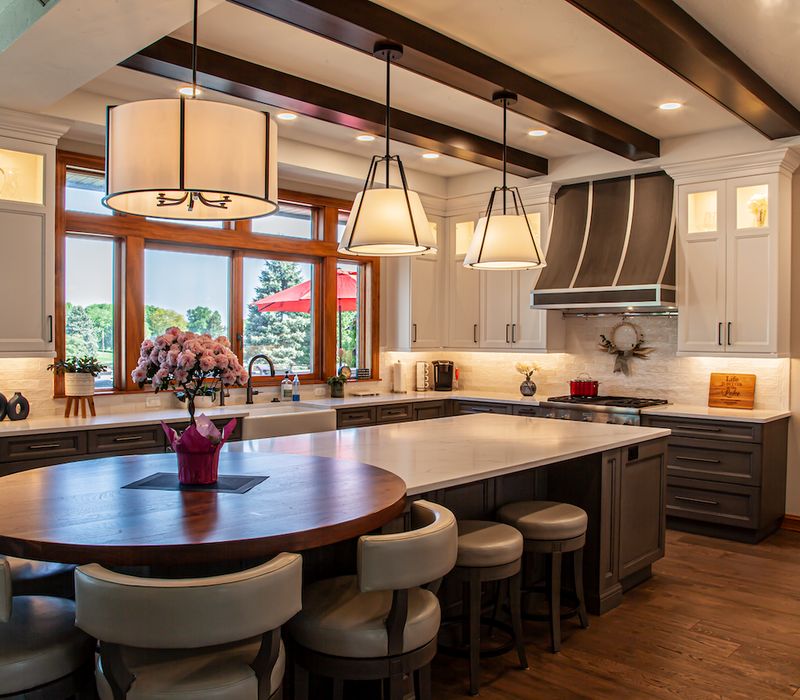 Gable end windows overlook the lake in this laid-back retreat featuring gray cabinetry, a black range hood with stainless steel accents, stone fireplaces, and wide plank wood floors. 