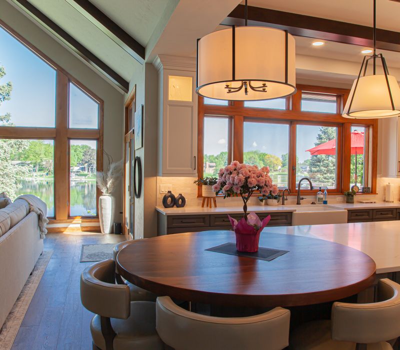 Gable end windows overlook the lake in this laid-back retreat featuring gray cabinetry, a black range hood with stainless steel accents, stone fireplaces, and wide plank wood floors. 