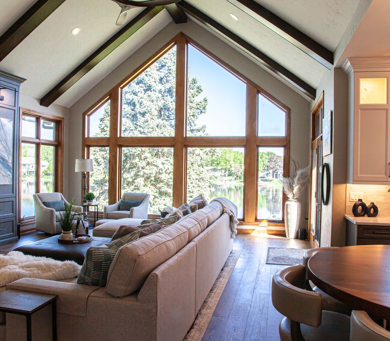Gable end windows overlook the lake in this laid-back retreat featuring gray cabinetry, a black range hood with stainless steel accents, stone fireplaces, and wide plank wood floors. 