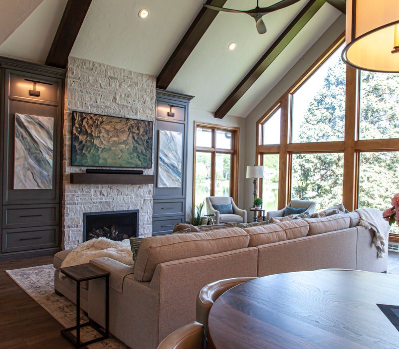 Gable end windows overlook the lake in this laid-back retreat featuring gray cabinetry, a black range hood with stainless steel accents, stone fireplaces, and wide plank wood floors. 