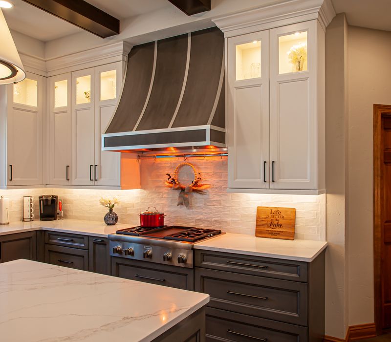 Gable end windows overlook the lake in this laid-back retreat featuring gray cabinetry, a black range hood with stainless steel accents, stone fireplaces, and wide plank wood floors. 