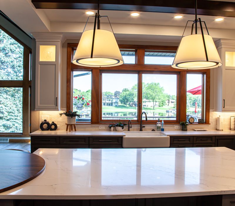 Gable end windows overlook the lake in this laid-back retreat featuring gray cabinetry, a black range hood with stainless steel accents, stone fireplaces, and wide plank wood floors. 