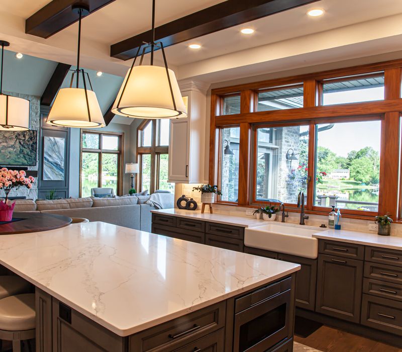 Gable end windows overlook the lake in this laid-back retreat featuring gray cabinetry, a black range hood with stainless steel accents, stone fireplaces, and wide plank wood floors. 