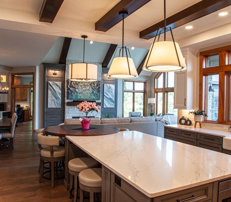 Gable end windows overlook the lake in this laid-back retreat featuring gray cabinetry, a black range hood with stainless steel accents, stone fireplaces, and wide plank wood floors. 