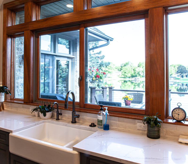 Gable end windows overlook the lake in this laid-back retreat featuring gray cabinetry, a black range hood with stainless steel accents, stone fireplaces, and wide plank wood floors. 