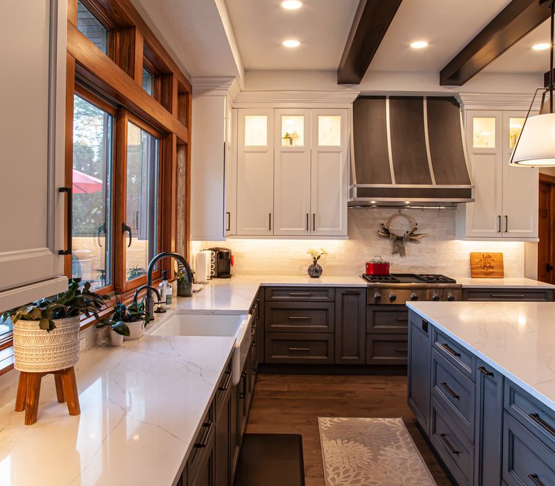 Gable end windows overlook the lake in this laid-back retreat featuring gray cabinetry, a black range hood with stainless steel accents, stone fireplaces, and wide plank wood floors. 