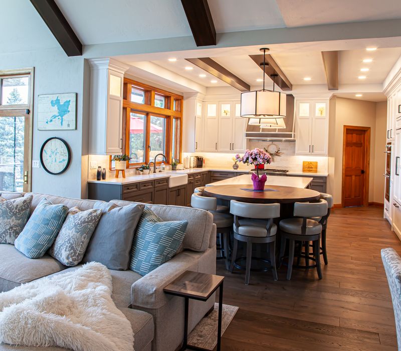 Gable end windows overlook the lake in this laid-back retreat featuring gray cabinetry, a black range hood with stainless steel accents, stone fireplaces, and wide plank wood floors. 