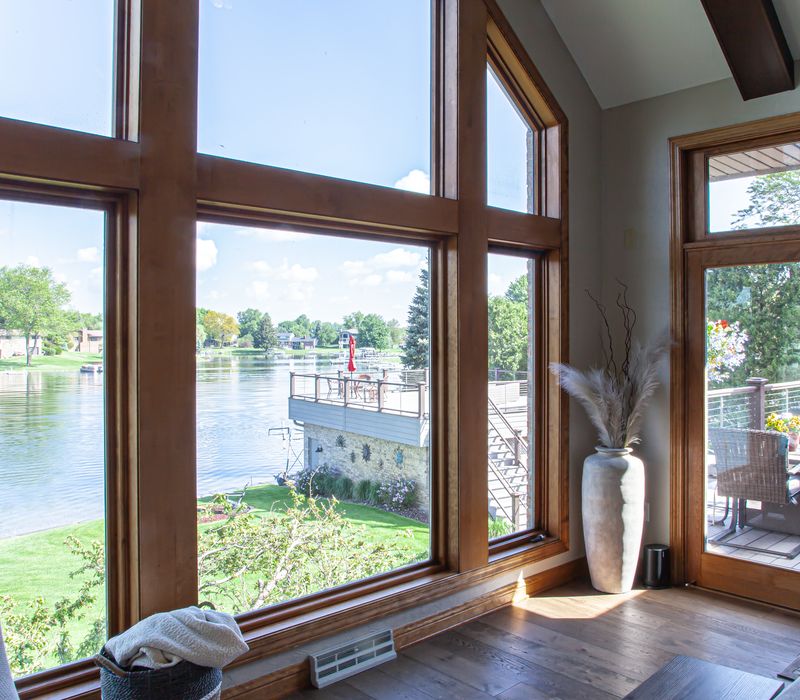 Gable end windows overlook the lake in this laid-back retreat featuring gray cabinetry, a black range hood with stainless steel accents, stone fireplaces, and wide plank wood floors. 