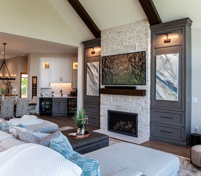 Gable end windows overlook the lake in this laid-back retreat featuring gray cabinetry, a black range hood with stainless steel accents, stone fireplaces, and wide plank wood floors. 
