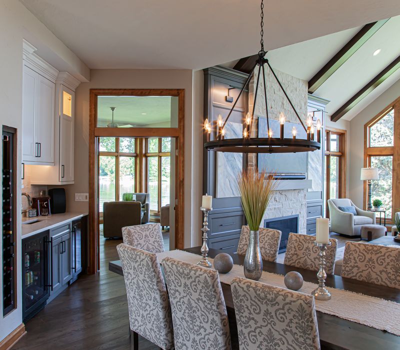 Gable end windows overlook the lake in this laid-back retreat featuring gray cabinetry, a black range hood with stainless steel accents, stone fireplaces, and wide plank wood floors. 