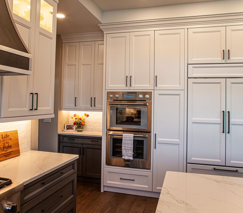 Gable end windows overlook the lake in this laid-back retreat featuring gray cabinetry, a black range hood with stainless steel accents, stone fireplaces, and wide plank wood floors. 
