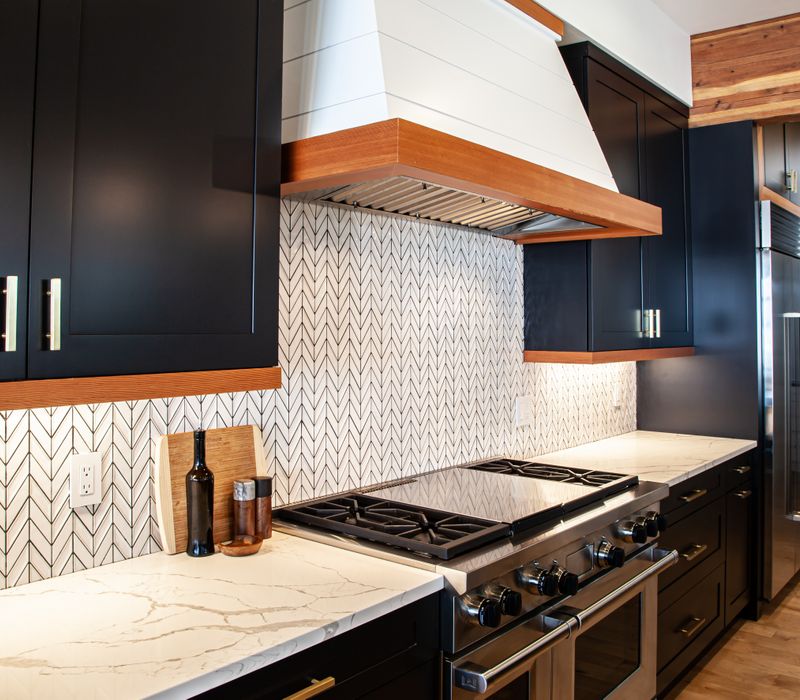 This mid-century modern home includes dramatic central stringer stairs behind a glass wall, white chevron tiles with dark grout, retro furniture, brushed gold fixtures, black cabinets, and laminated beams. 