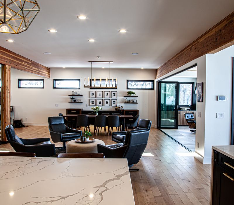 This mid-century modern home includes dramatic central stringer stairs behind a glass wall, white chevron tiles with dark grout, retro furniture, brushed gold fixtures, black cabinets, and laminated beams. 