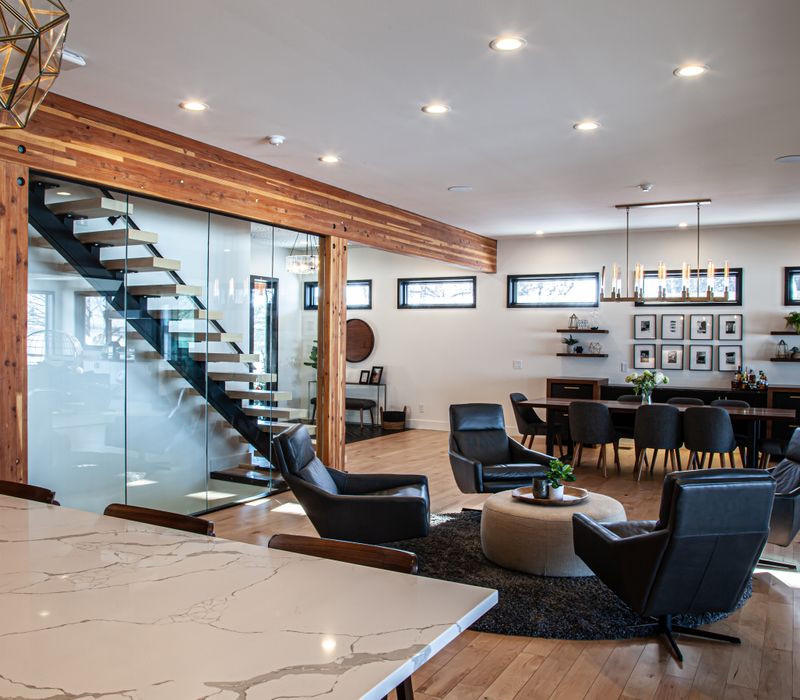 This mid-century modern home includes dramatic central stringer stairs behind a glass wall, white chevron tiles with dark grout, retro furniture, brushed gold fixtures, black cabinets, and laminated beams. 