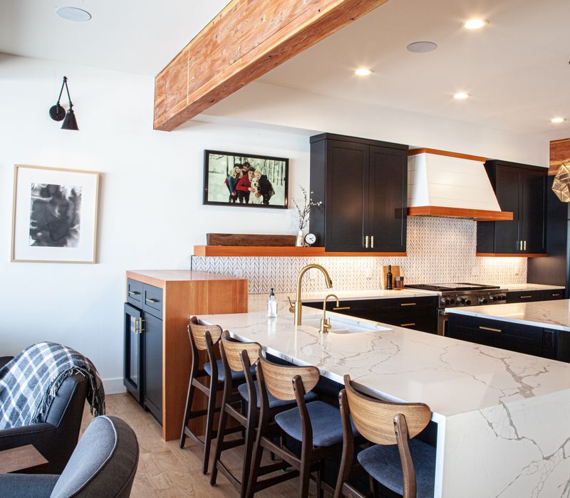 This mid-century modern home includes dramatic central stringer stairs behind a glass wall, white chevron tiles with dark grout, retro furniture, brushed gold fixtures, black cabinets, and laminated beams. 