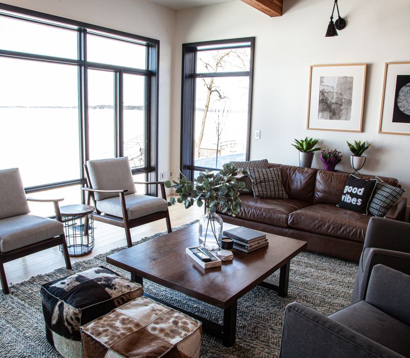 This mid-century modern home includes dramatic central stringer stairs behind a glass wall, white chevron tiles with dark grout, retro furniture, brushed gold fixtures, black cabinets, and laminated beams. 