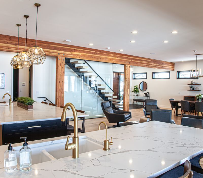 This mid-century modern home includes dramatic central stringer stairs behind a glass wall, white chevron tiles with dark grout, retro furniture, brushed gold fixtures, black cabinets, and laminated beams. 