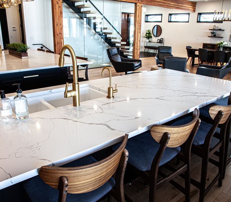 This mid-century modern home includes dramatic central stringer stairs behind a glass wall, white chevron tiles with dark grout, retro furniture, brushed gold fixtures, black cabinets, and laminated beams. 